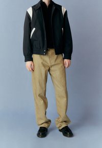 Czarna kurtka typu bomber z kremowymi akcentami, beżowe spodnie i czarne buty. Kurtka ma elastyczne ściągacze na rękawach, kieszenie oraz usztywniony kołnierz.