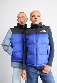 Blauwe puffer vest met zwarte bovenkant, voorzien van een ritssluiting en hoge kraag. De stof is glad met een gewatteerde textuur.