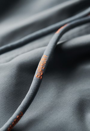 Szara tkanina z pomarańczowym logo "Superdry". Tekstura wydaje się gładka, a szczegóły obejmują sznurowadło o teksturowanym, wzorzystym wyglądzie.