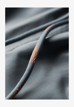 Szara tkanina z pomarańczowym logo "Superdry". Tekstura wydaje się gładka, a szczegóły obejmują sznurowadło o teksturowanym, wzorzystym wyglądzie.