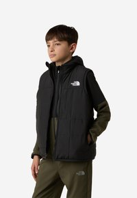 Fekete, kapucnis bélelt mellény, cipzáros záródással és fehér logóval a bal mellkason, fekete és olívazöld ruházat fölött viselve.