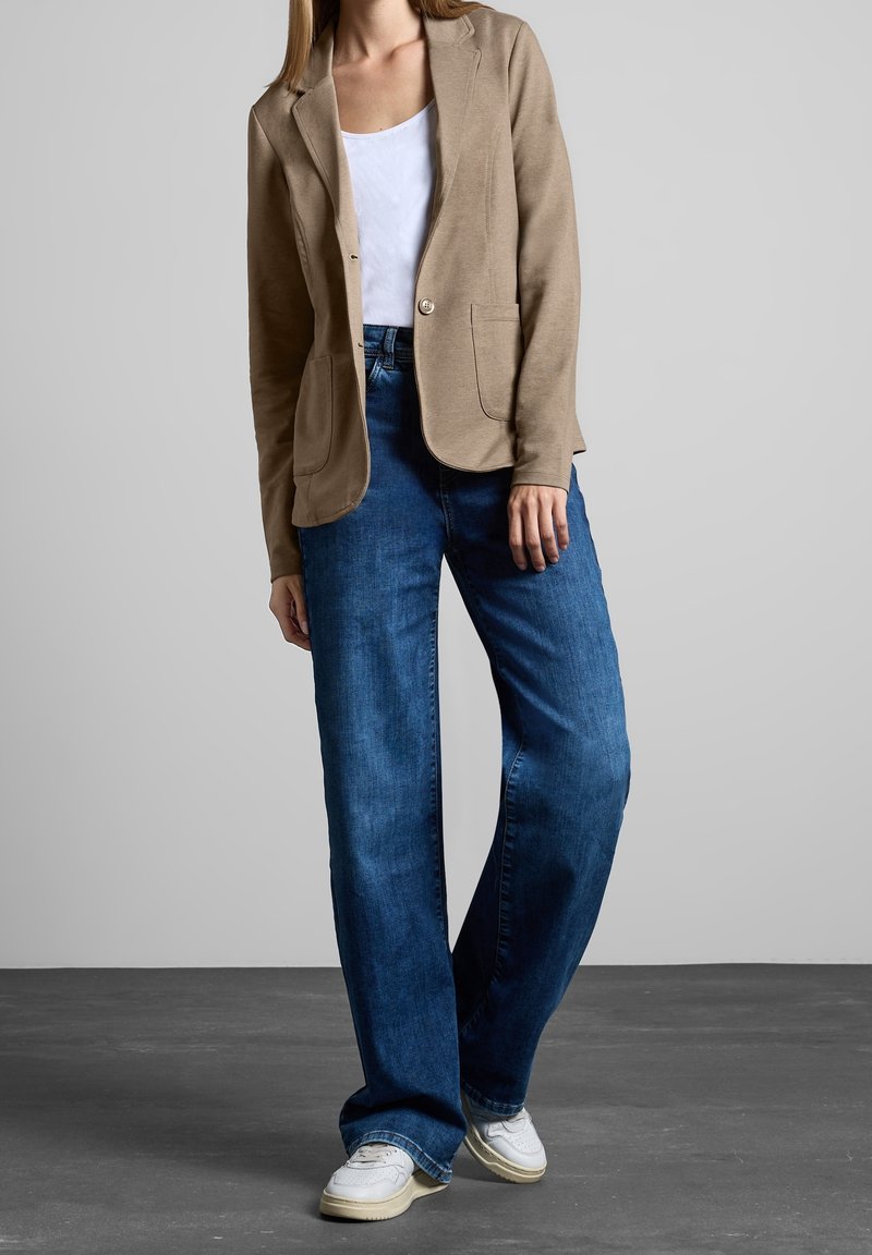 Femme portant un blazer beige sur un haut blanc, associé à un jean bleu ample et des baskets blanches, debout devant un fond gris uni.