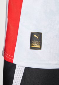 Červený a biely futbalový dres vyrobený z texturovanej tkaniny; má čiernu a zlatú nálepku s nápisom "AUTORIZOVANÝ LICENCOVANÝ FUTBAL."