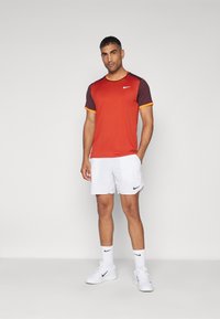 Miestenska súprava športového oblečenia obsahujúca červené a mahagónové tričko s krátkym rukávom s oranžovými akcentmi a biele šortky s čiernym logom Nike.