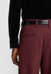 Bordeauxrote Hose mit strukturierter Stoffqualität und schwarzem Ledergürtel, ausgestattet mit Seitentaschen und geradem Schnitt. Schwarzes Samthemd mit Knöpfen.