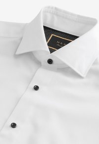 White formal shirt with black buttons and a pointed collar, showing a black label with gold trim inside the collar.