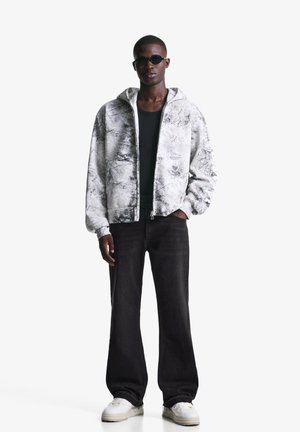 Giacca con cappuccio grigia con pattern nero marmorizzato, abbinata a una maglietta nera, jeans scuri e sneakers bianche; occhiali da sole neri rotondi.
