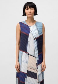 Mouwloze jurk met een gemengd geometrisch patroon in blauw, marineblauw en beige, voorzien van een geplooide textuur en een knoopdetail bij de halslijn.