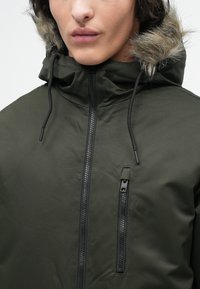 Olivengrøn jakke med tekstureret finish, der har en faux fur-fortykket hætte og to frontlomme med lynlås, detaljeret med sorte lynlåse og træk.