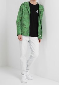 Zelená maskáčová bunda s kapucí, nošená přes černé tričko, v kombinaci s bílými džíny a bílými teniskami. Hladká, lehká tkanina.