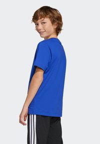 Blue cotton T-shirt with a crew neck and short sleeves. The back features a clean design. Worn with black pants that have white stripes.