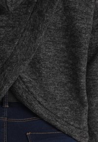 Węgielnoszary sweter z dzianiny o kopertowym kroju, z miękką teksturą i drapowanym dekoltem, zestawiony z ciemnoniebieskimi dżinsami z kontrastującym przeszyciem.