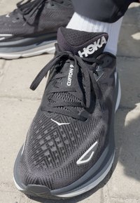 Scarpa da corsa nera con tessuto in rete, motivo testurizzato, collare imbottito, dettagli riflettenti e un logo bianco sulla linguetta.