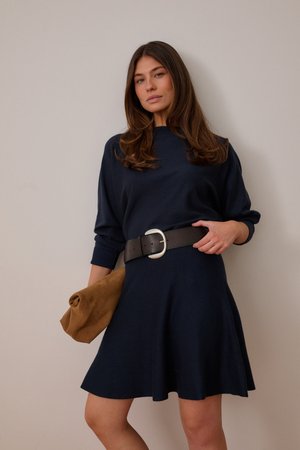Žena v námorníckych modrých šatách so širokým čiernym opaskom drží hnedú kabelku na jednofarebnom béžovom pozadí.