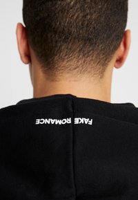 Black hooded sweatshirt featuring a stitched white label reading "FAKE ROMANANCE" on the back seam. Soft cotton material.