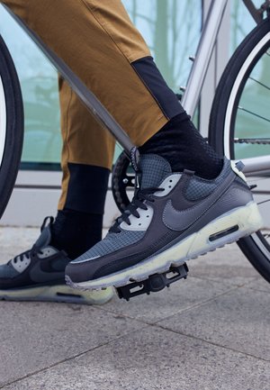 Personne portant des baskets grises et noires avec des chaussettes noires et un pantalon marron, pied sur une pédale de vélo près d'une roue de vélo grise.