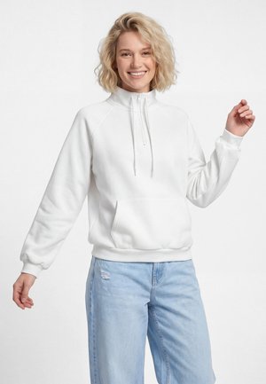 Donna sorridente con capelli biondi e ricci che indossa una felpa bianca con zip e jeans azzurri strappati su sfondo bianco.