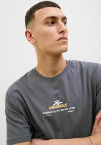 Harmaa puuvillapaita, jossa on graafinen printti palmun kuvasta. Tekstissä lukee "ORIGINALS" ja "Crafted by the denim crew" erottuvissa väreissä.