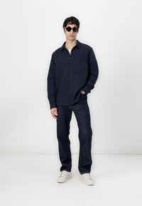 Navy button-up shirt with a chest pocket, paired with dark jeans and white sneakers. Model wears round sunglasses, standing against a white backdrop.