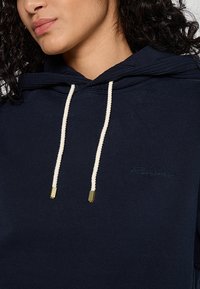 Hoodie bleu marine en tissu doux, avec un cordon de serrage agrémente d'accents dorés et un logo ton sur ton brodé sur la poitrine.
