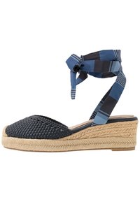 Klínové espadrilky z tmavě modré tkané látky s podešví z copánkového juty. Mají modrou pruhovanou stuhu na zavazování kolem kotníku a zaoblený tvar špičky.