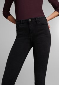 Jeans negros de cintura alta con un diseño ajustado, que cuentan con un cierre de botón metálico y detalles de costura sutiles en los bolsillos. Textura suave.