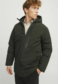 Chaqueta acolchada verde oscura con capucha, que cuenta con un bolsillo en el pecho con cremallera y dos bolsillos frontales, fabricada en un material suave y resistente al agua.