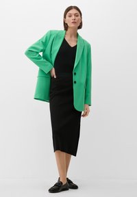 Zeleno blazer z izrezanim ovratnikom in dvema črnima gumboma, v kombinaciji s črno prilegajočo krilom in črnimi loaferji, v beli barvi ozadja.