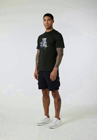 Camiseta de algodón negra con un gráfico de oso y detalles en verde, combinada con pantalones cortos de cargo azul marino y zapatillas blancas, mostrando un ajuste informal.