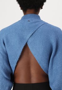 Maglione blu a costine con un design a schiena aperta. Presenta un colletto alto e un dettaglio del logo visibile vicino al collo. Tessuto morbido.