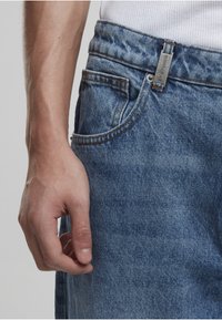 Zbliżenie na dłoń osoby obok przedniej kieszeni niebieskich dżinsów z metalową metką na szlufce paska.