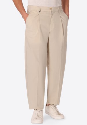 Pantaloni beige plissettati con tasche frontali indossati con sneakers bianche e mani infilate, mostrati dalla vita ai piedi su sfondo chiaro.