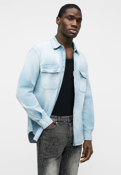 Άνδρας που φοράει ανοιχτό μπλε πουκάμισο τζιν πάνω από μαύρο τοπ και σκισμένα σκούρα γκρι τζιν, στέκεται με το ένα χέρι στην τσέπη μπροστά σε λευκό φόντο.