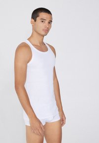 Débardeur en coton blanc avec un col rond, sans manches et de coupe ajustée. Porté avec un slip blanc assorti, sur fond neutre.