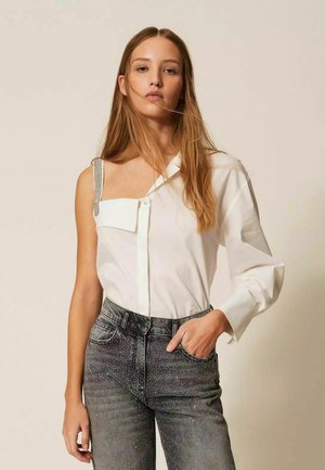 Giovane donna con capelli lunghi che indossa una camicetta bianca asimmetrica con una manica scoperta e jeans scuri scintillanti, mano in tasca.