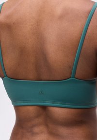 Οπίσθια όψη ενός ατόμου που φοράει ένα τιρκουάζ αθλητικό σουτιέν με λεπτές τιράντες, δείχνοντας το πάνω και μεσαίο μέρος της πλάτης με δέρμα και μυϊκό τόνο.