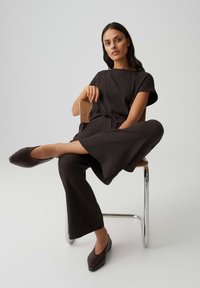 Woman in dark textured outfit and pointed flats sits cross-legged on wooden chair with metal legs against plain background.