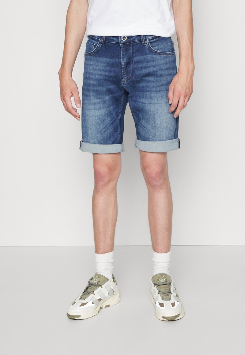 Shorts in denim di blu medio con un lavaggio sbiadito e risvolti. Indossati con calzini bianchi e sneaker chiare con dettagli verdi.