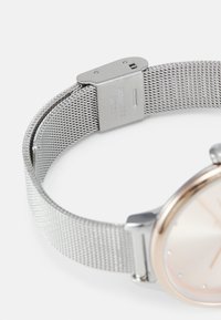 Reloj con correa de malla plateada y caja redonda en tono oro rosado, con una esfera en color melocotón claro. Presenta marcadores de horas minimalistas y cierre de presión.