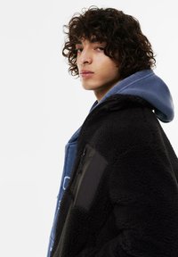 Czarna polarowa kurtka z gładką, ciemną nylonową kieszenią i niebieską bluzą z kapturem pod spodem. Kręcone włosy częściowo zasłaniają twarz.