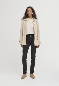 Abrigo beige sobre un suéter claro, combinado con jeans negros y mocasines beige. Presenta un cierre de cinco botones en los jeans y cabello rizado.