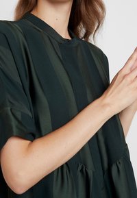Abito verde scuro con maniche corte, caratterizzato da strisce verticali di satin, un collo rotondo e una vita arricciata per una silhouette fluida.