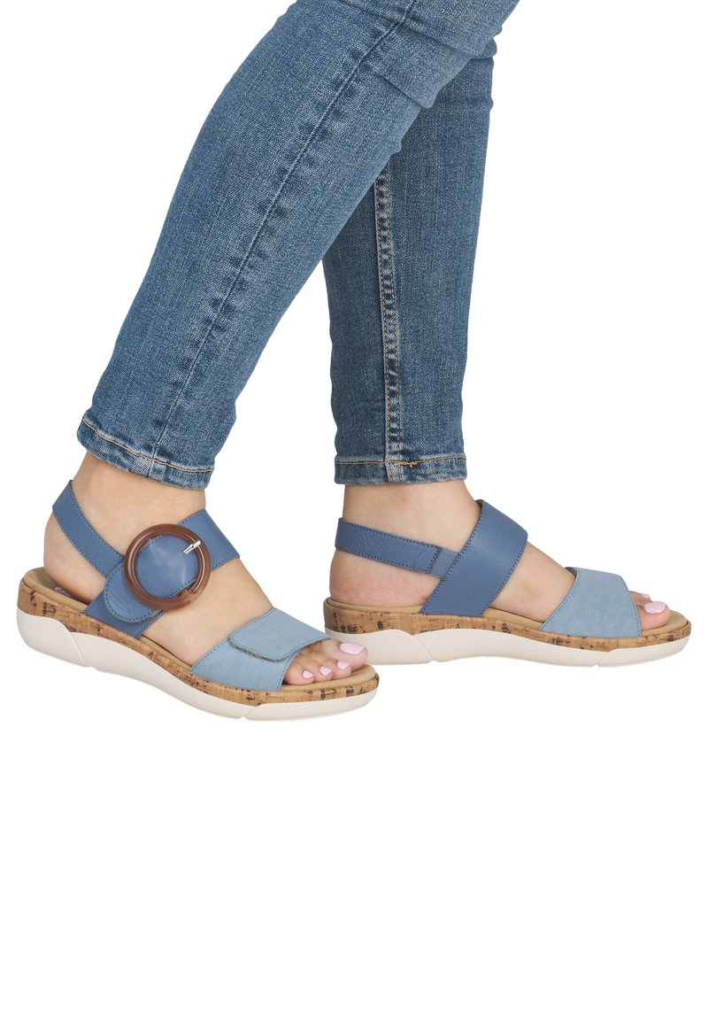 Blaue Sandalen mit Korksohlen, flachem Design, breiten Riemen und einem großen runden Akzent. Glatte Textur und offene Zehenpartie.