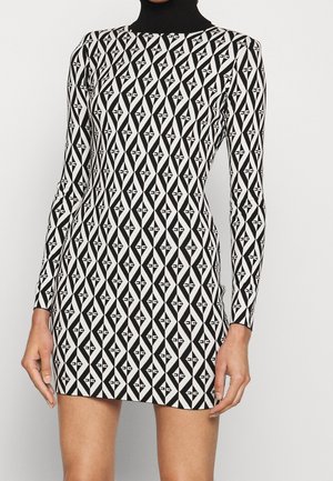 Vestido mini ajustado con estampado geométrico en blanco y negro, mangas largas y cuello alto negro, llevado por una persona de pie.