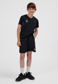 Completo sportivo nero composto da maglietta e pantaloncini, realizzato in materiale leggero. Presenta dettagli con logo, motivi laterali e vestibilità classica. Indossato con scarpe da ginnastica nere e calze bianche.