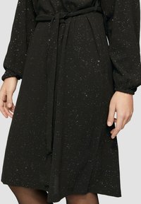 Schwarzes Kleid mit funkelnder Textur, langen Ärmeln und tailliertem Bund. Es hat einen knielangen Saum und ein dezentes, schimmerndes Muster überall.