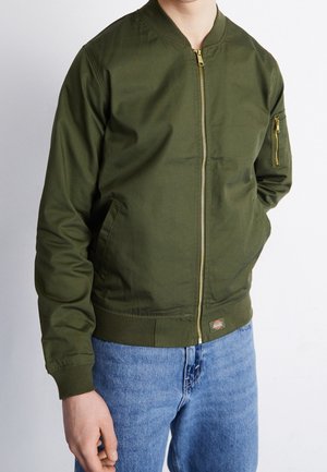 Personne portant une veste bomber zippée vert olive avec une poche zippée sur la manche et un jean bleu, mains dans les poches devant un fond uni.