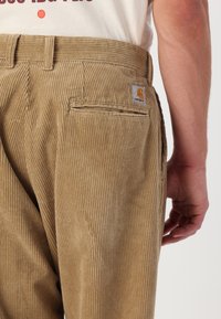 Pantalones de pana marrones con costuras verticales. Cuenta con un bolsillo trasero y un parche del logo de Carhartt en la cinturilla. Textura suave.