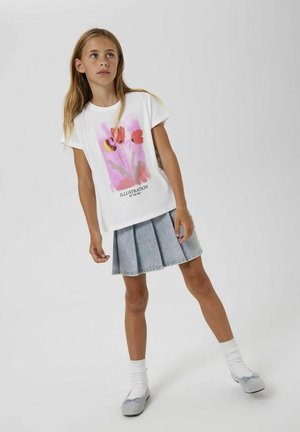 T-shirt blanc avec illustration florale rose et rouge, associé à une jupe plissée en denim clair. Le modèle porte des chaussettes blanches et des chaussures grises.