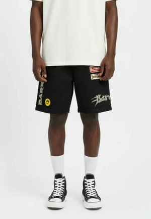 Mężczyzna ubrany w czarne szorty z nadrukiem, białe skarpetki sportowe, czarne sneakersy za kostkę i białą koszulkę, stojący na tle jednolitego tła.